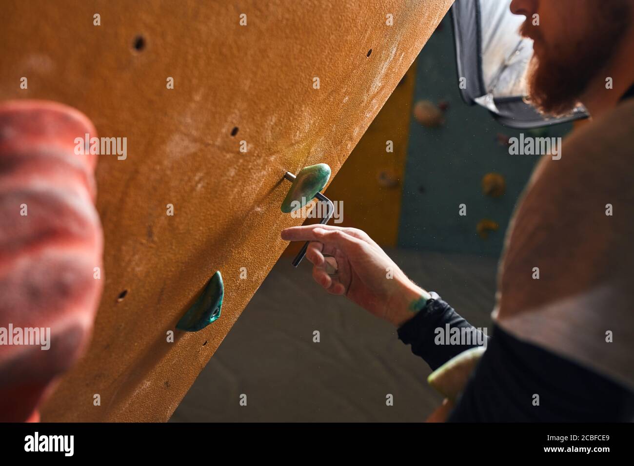 Vue rapprochée d'un jeune grimpeur méconnaissable avec barbe qui fixe la roche verte artificielle au mur d'escalade jaune, vérifie les cales et les panneaux avant la formation Banque D'Images