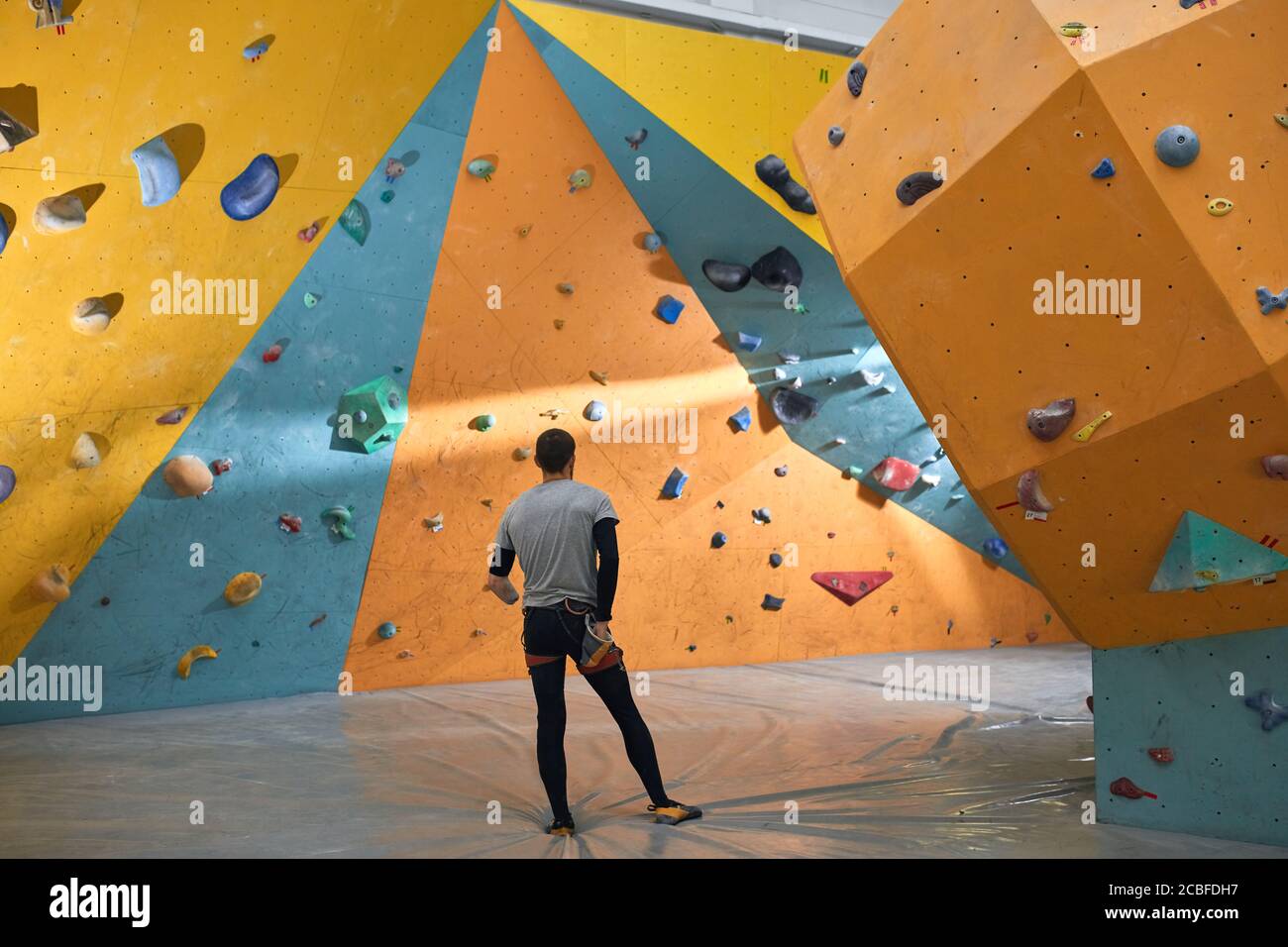Jeune homme boulderer avec handicap physique debout avec soin dans la salle de gym d'escalade, regardant les murs de roche artificielle colorée, en choisissant où pour le stament Banque D'Images