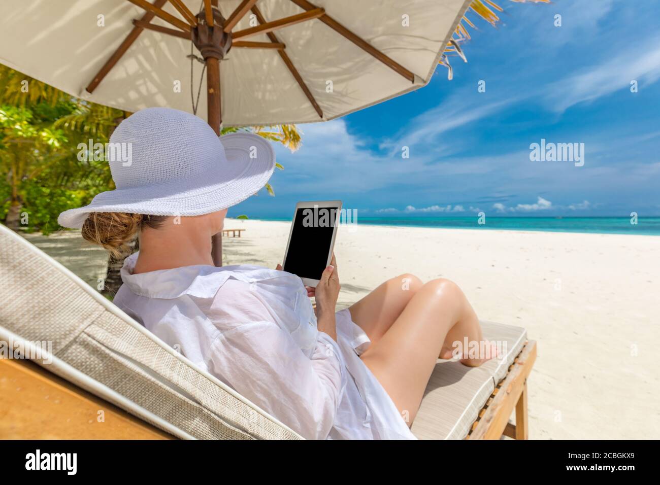 Femme avec une tablette sur la plage assise dans la chaise longue. Jeune femme chapeau avec tablette pc à la plage, vacances d'été de luxe, loisirs Banque D'Images