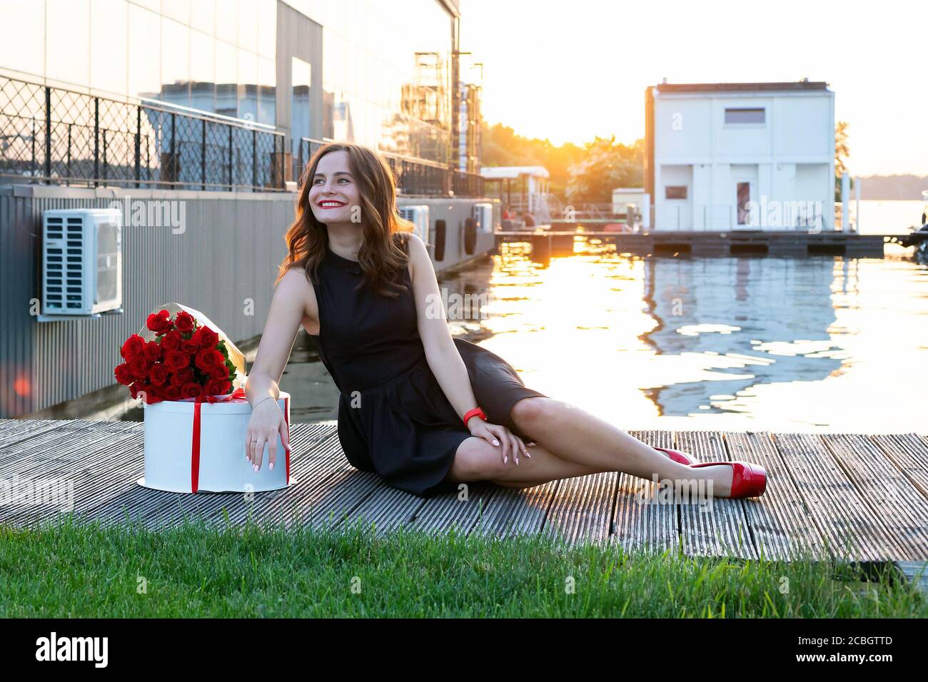 Fille D Anniversaire Souriante Avec Cheveux Bruns Dans Une Petite Robe Noire Assise Sur Le Sentier En Bois Au Coucher Du Soleil Boite Ronde Avec Gateau D Anniversaire Enveloppe De Rouge Ri Photo Stock
