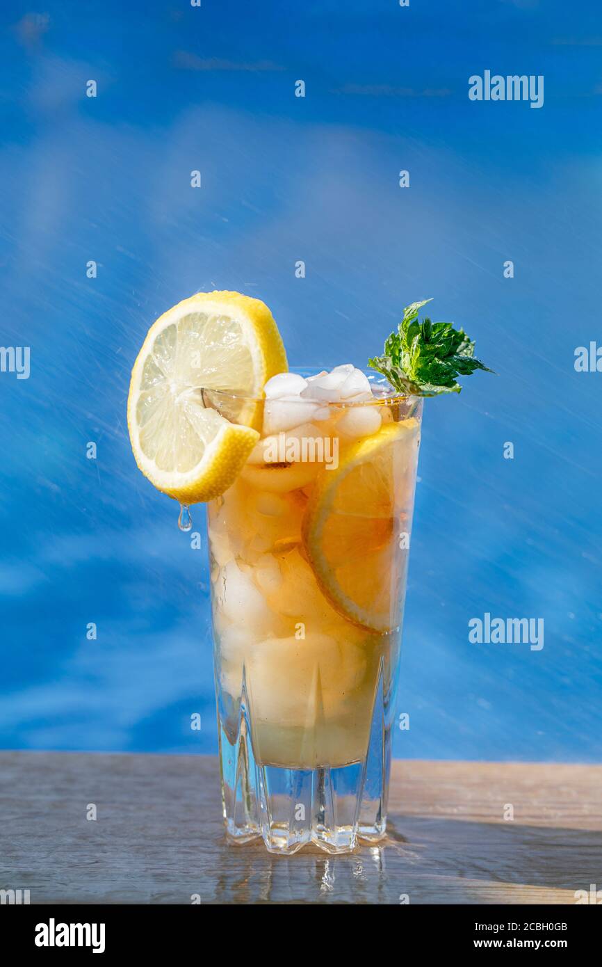 Un verre de thé glacé avec citron et menthe, piscine avec eau bleue en arrière-plan. Image verticale. Il y a un citron tranché sur le bord du verre a Banque D'Images