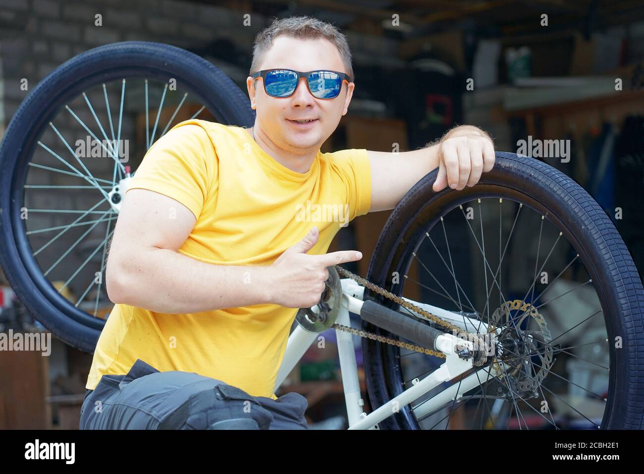 un gars avec des lunettes et un t-shirt jaune est assis avant d'une bicyclette sur le fond d'un atelier garage Banque D'Images
