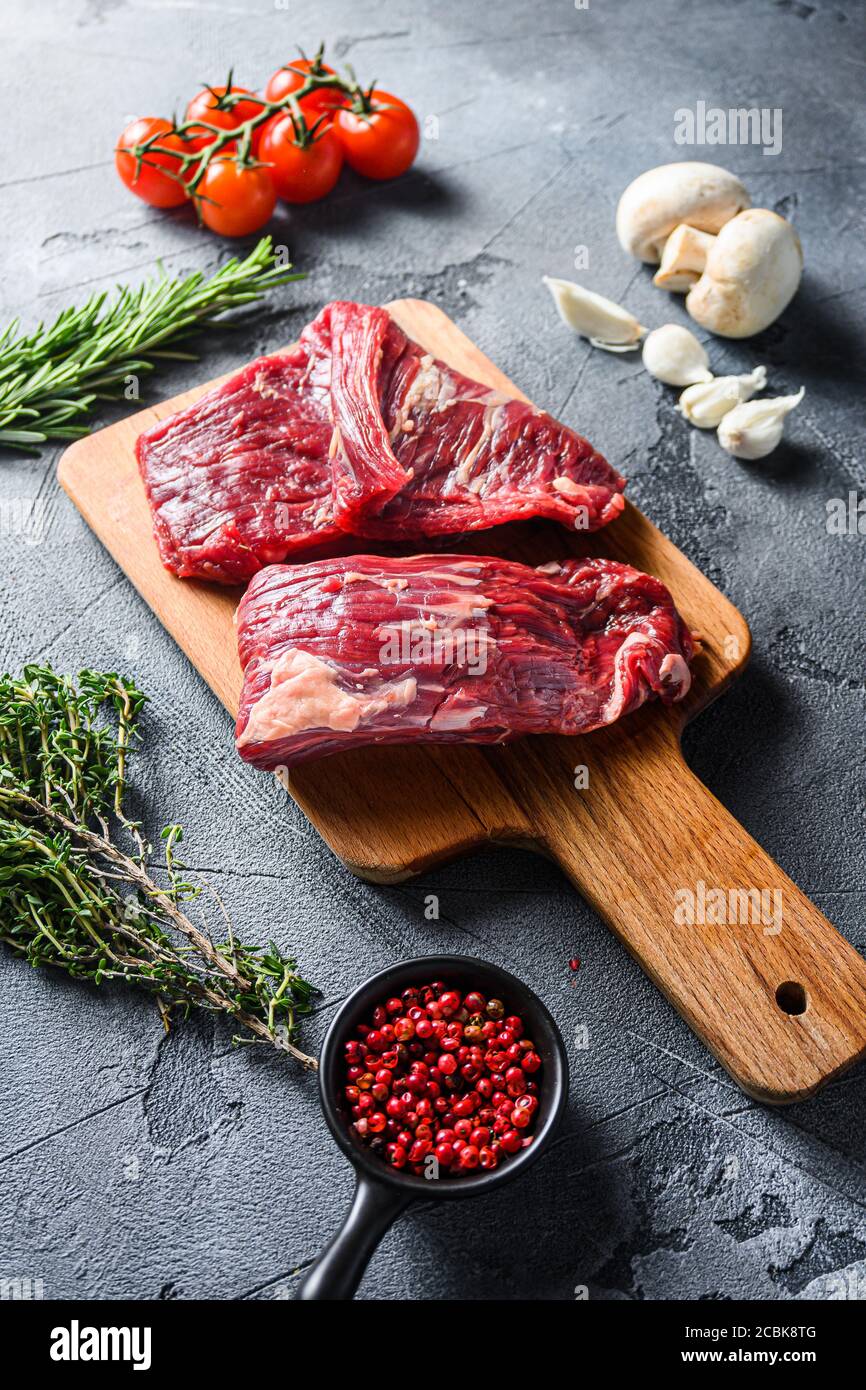 Steak à rabat cru coupé avec machette, steak de jupe, sur planche à découper en bois, avec tomates fines poivrières sur fond de pierre grise Banque D'Images