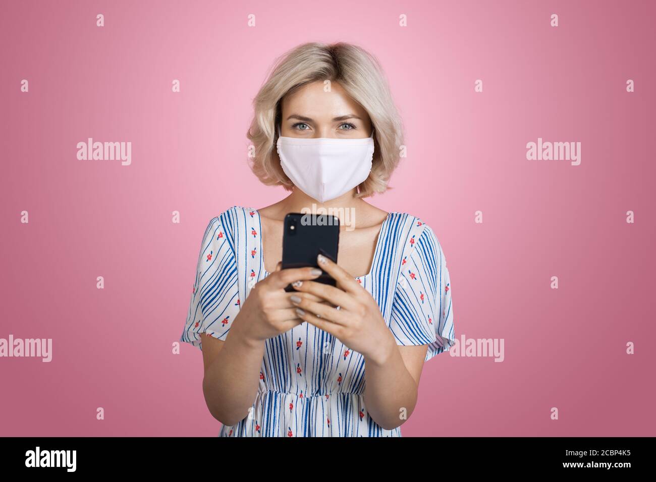 Jolie jeune femme blonde portant une robe et un masque médical est en train de discuter sur mobile en souriant à l'appareil photo sur un rose mur du studio Banque D'Images