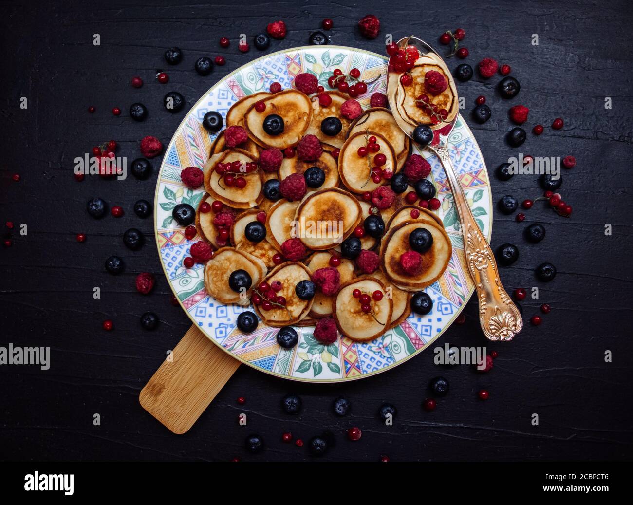 Mini crêpes aux baies sur une assiette à motifs avec une cuillère en argent de style rustique. Petit déjeuner traditionnel sur table noire Banque D'Images