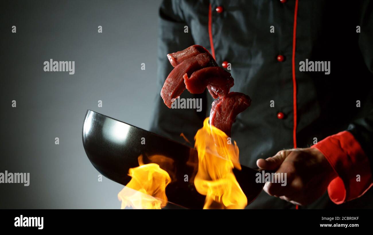 Gros plan sur le chef qui lance des steaks de bœuf crus dans un poêle à wok au feu. Préparation de plats asiatiques frais sur fond sombre. Banque D'Images