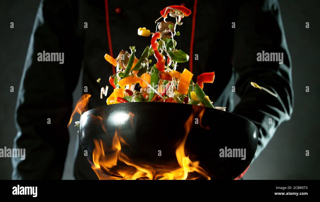 Gros plan du chef qui lance le mélange de légumes dans un poêle à wok au feu. Préparation de plats asiatiques frais sur fond sombre. Banque D'Images