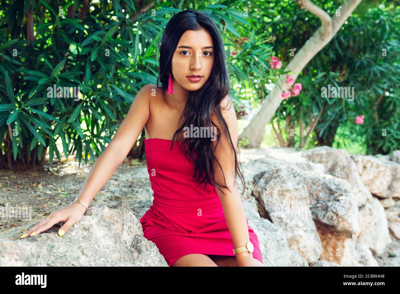 Jeune fille latine avec de longs cheveux noirs et robe rouge, dans une zone de végétation et de rochers. Banque D'Images