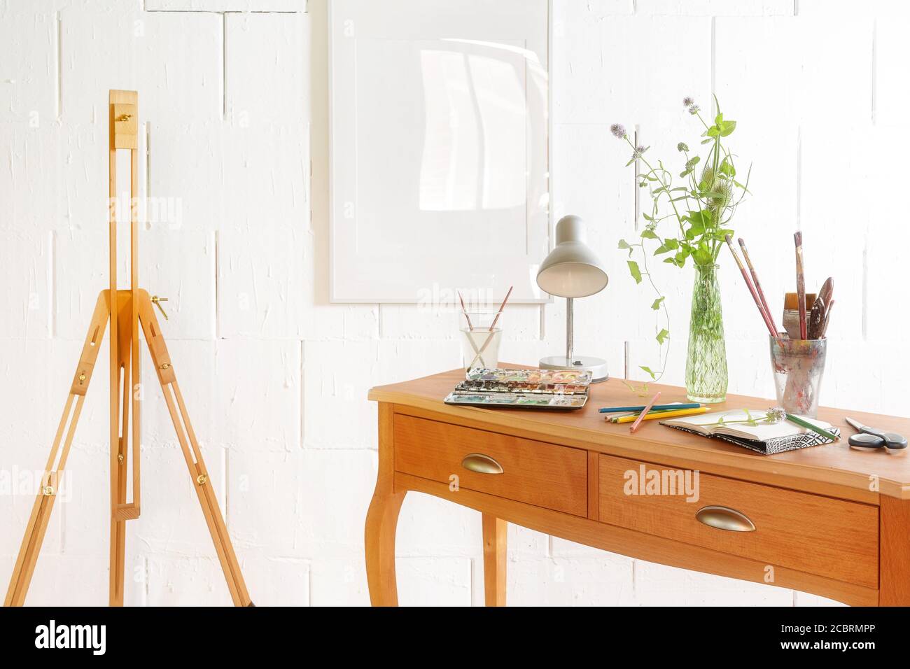 Petit espace pour être créatif, crayons, carnet d'esquisses, aquarelle et brosses sur une console de tiroir en bois et un chevalet vide contre un mur blanc rugueux, copie Banque D'Images