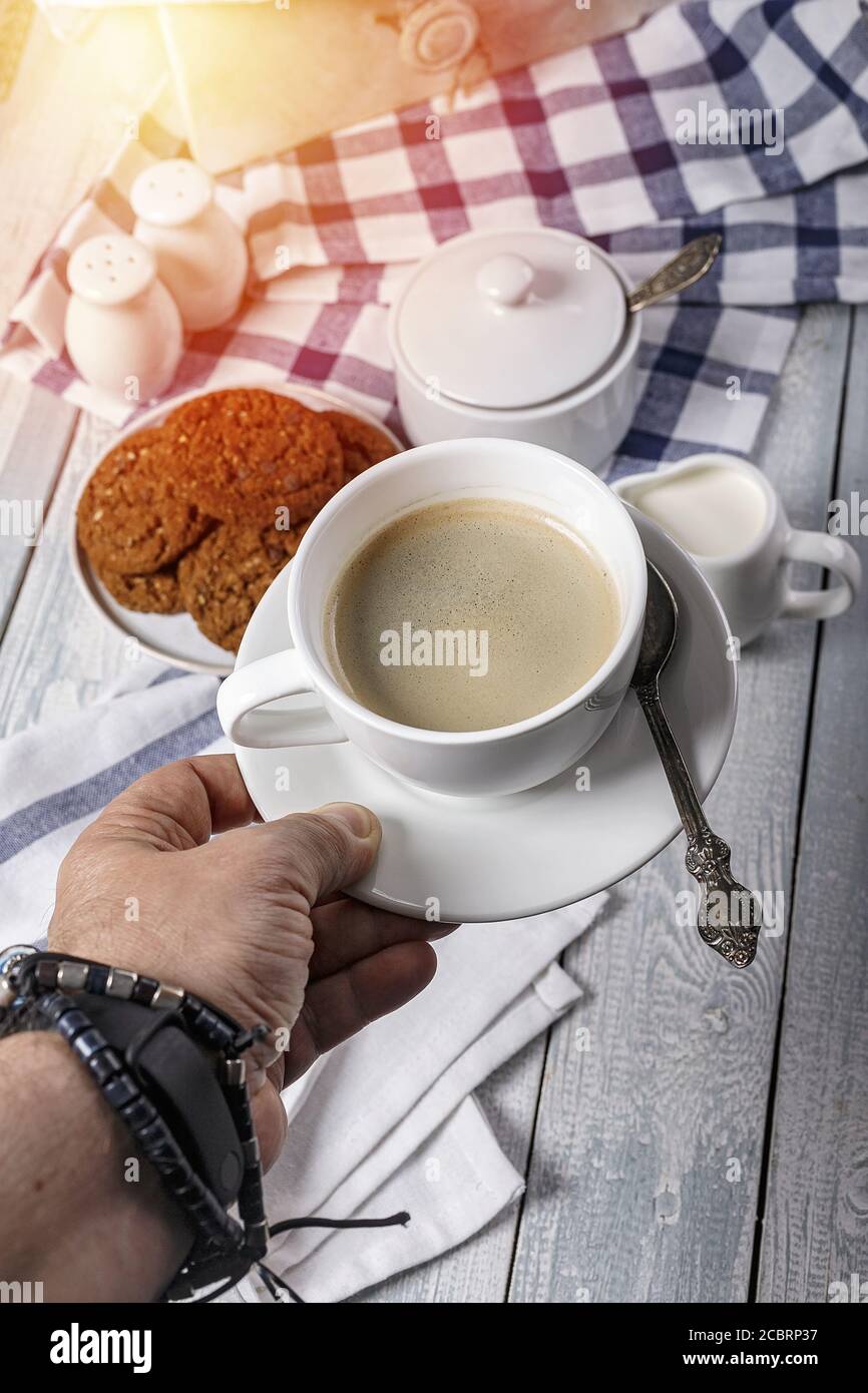 Main du Mans tenant une tasse de café blanc. Sur le fond d'une table en bois avec de la crème, un bol à sucre et des biscuits aux flocons d'avoine. Copier l'espace. Sho vertical Banque D'Images