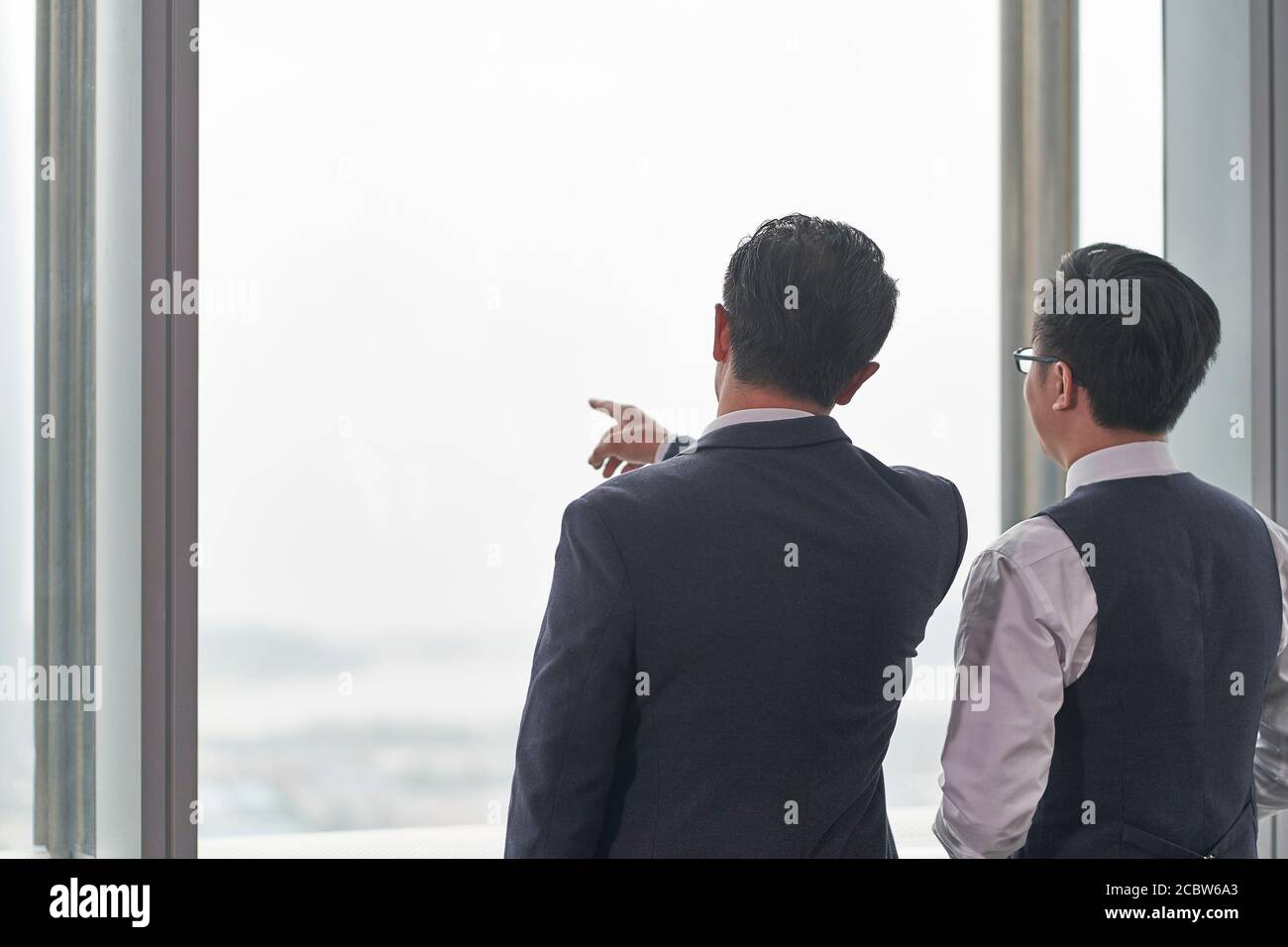 vue arrière de deux hommes d'affaires asiatiques debout devant de la fenêtre de bureau ayant une discussion Banque D'Images