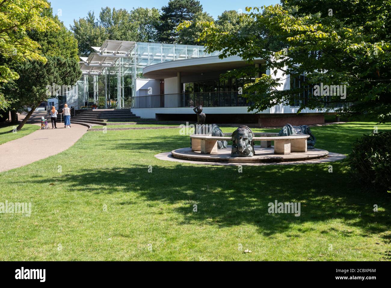 Adultes et enfants marchant dans les jardins Jephson en face de la Glasshouse avec la sculpture de siège Elephant Circle au premier plan. Banque D'Images