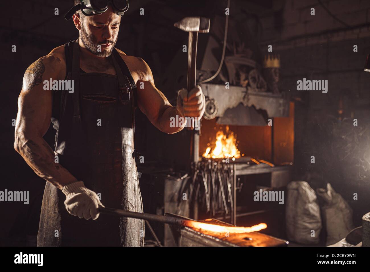 adulte musclé avec barbe portant un tablier en cuir travaillant dans l'atelier, chauffant. métal trempé dans le four Banque D'Images