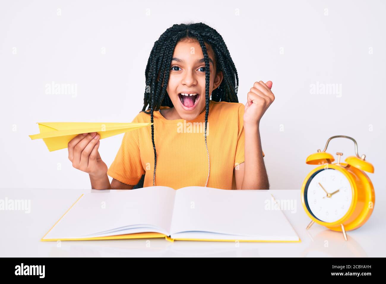 Jeune fille afro-américaine avec des tresses tenant un avion de papier tout en étudiant criant fier, célébrant la victoire et le succès très excité avec Banque D'Images