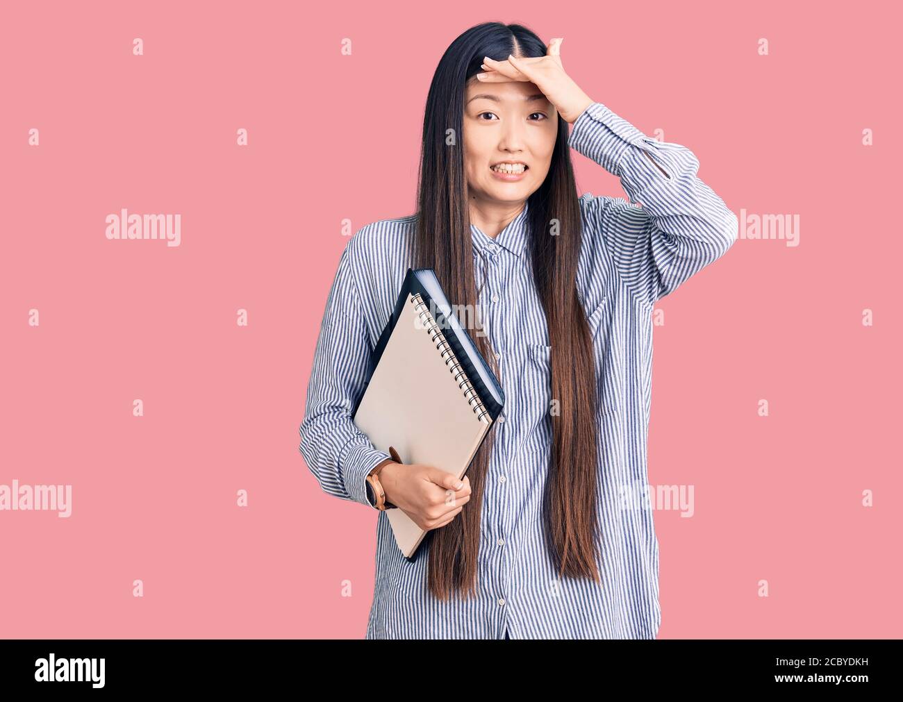 Jeune belle chinoise tenant le carnet stressé et frustré de la main sur la tête, surpris et fâché visage Banque D'Images
