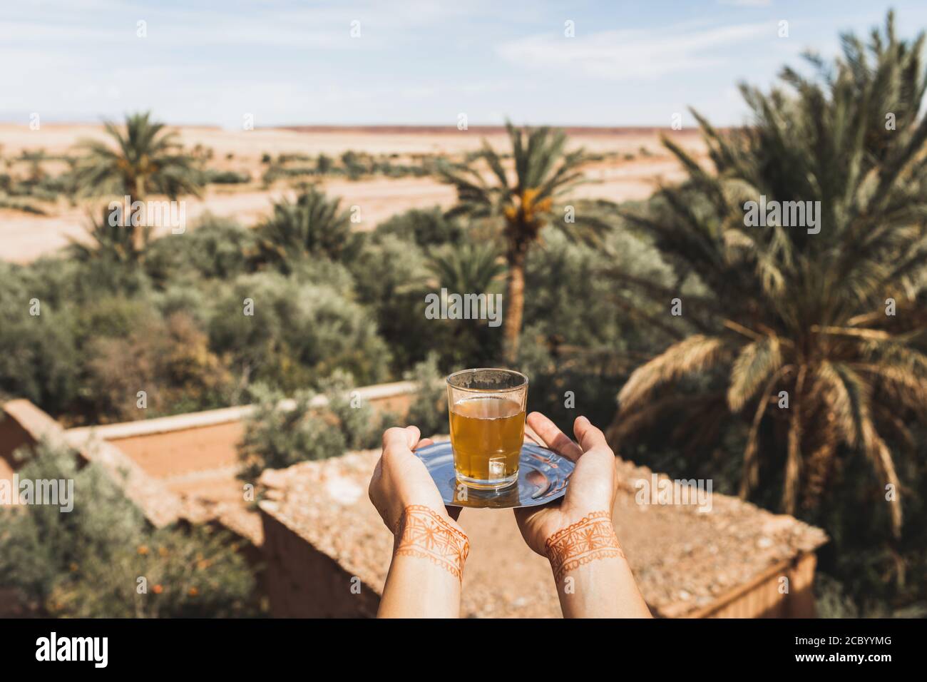 Femme mains avec tatouage au henné tenant un verre de thé marocain à la menthe traditionnel. Sable désert et palmiers sur fond. Banque D'Images
