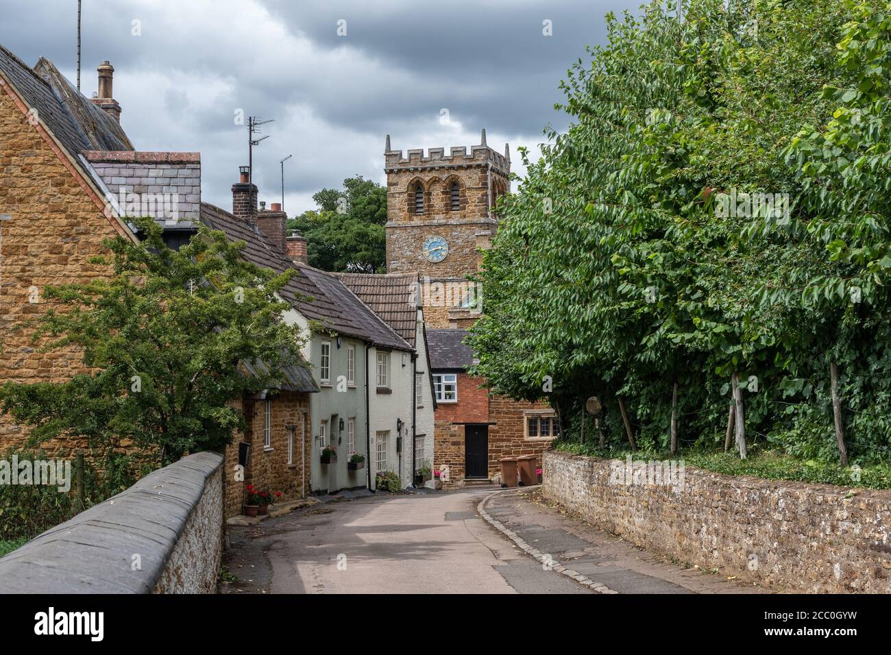 Scène de village traditionnelle, Mears Ashby, Northamptonshire, Royaume-Uni; route bordée d'arbres et de maisons et tour d'église au milieu de la distance. Banque D'Images