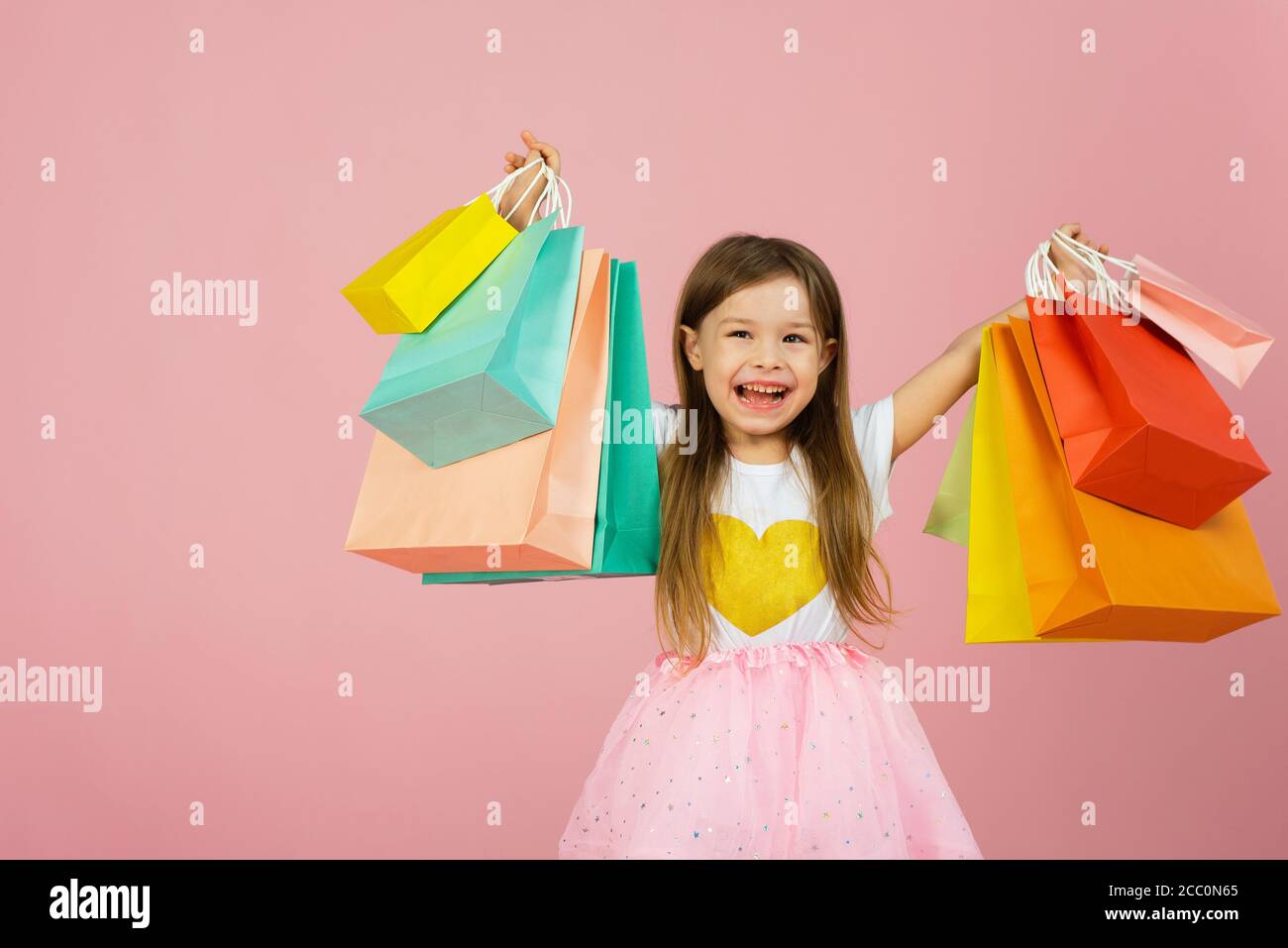 Petite fille avec de nombreux sacs de shopping sur le dos rose pastel. Jolie jeune fille joyeuse dans la jupe de tulipe, avec de longs cheveux blonds marchant avec des paquets colorés sur fond rose. Banque D'Images