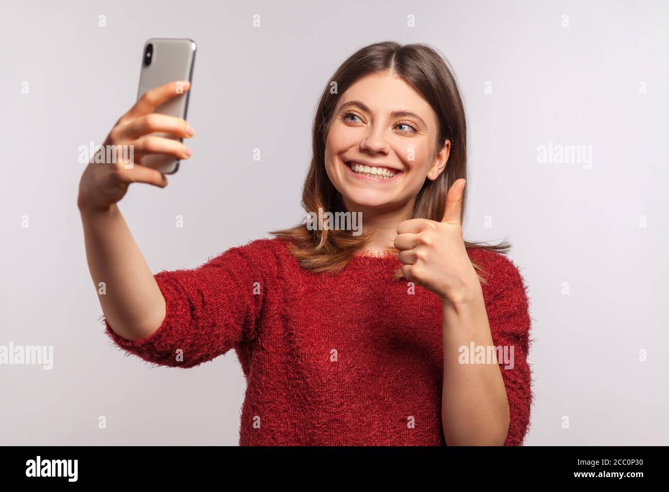 Portrait d'une petite fille sympathique dans un chandail déchiqueteur faisant un appel vidéo par téléphone mobile et montrant le pouce vers le haut, communiquant à l'aide d'Internet-activé électronique Banque D'Images