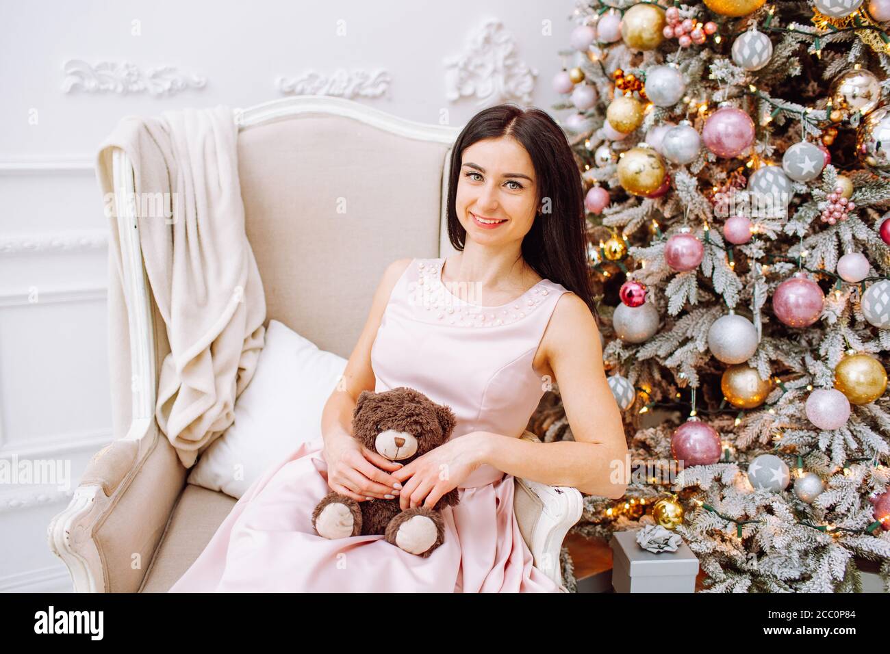 Femme en robe rose avec petit ours assis sur une chaise près d'un arbre de Noël clair. Nouvel an, Noël, vacances Banque D'Images