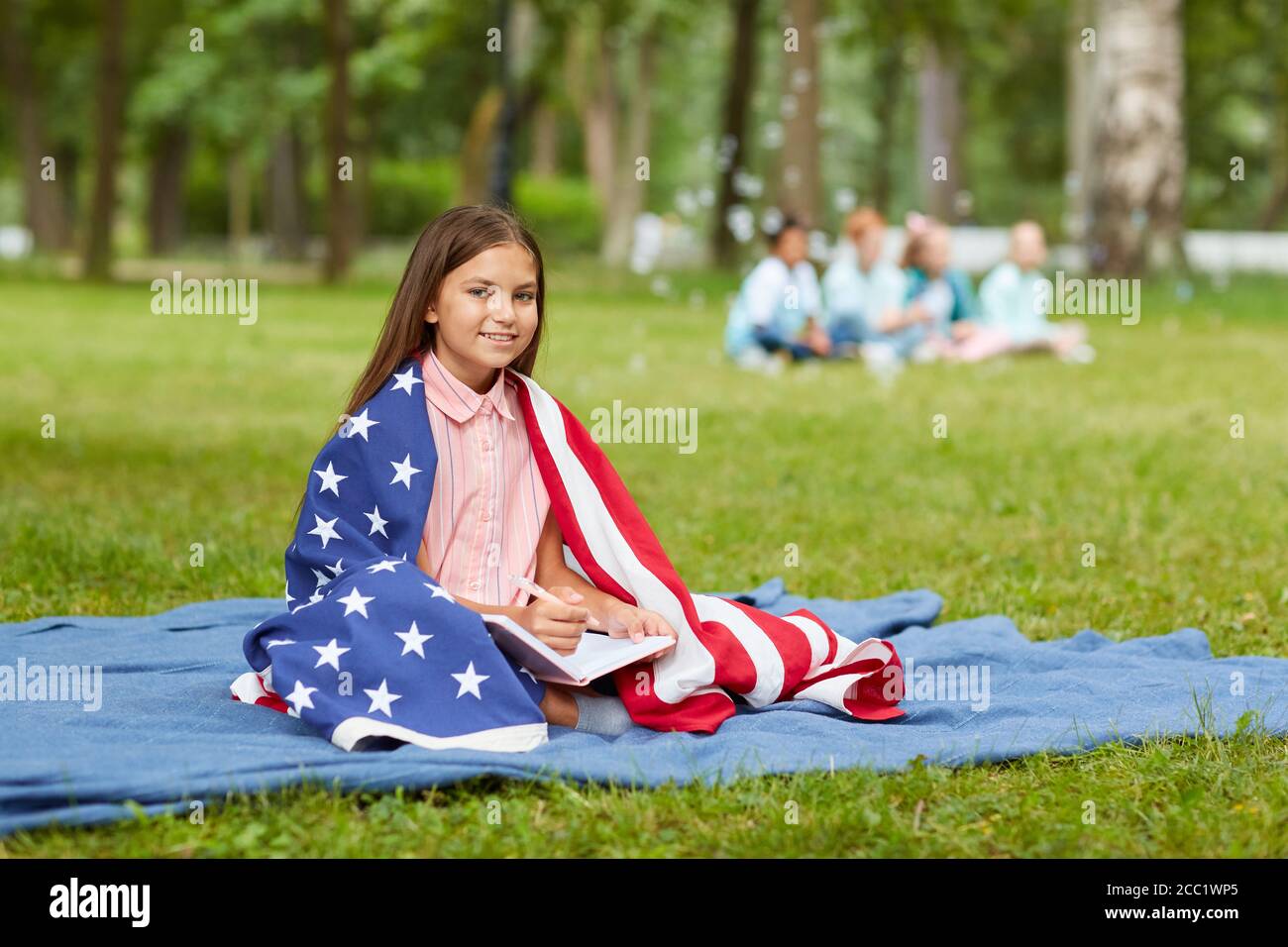 Portrait en longueur de petite fille couverte par un drapeau américain, assis sur une couverture de pique-nique dans le parc et souriant à l'appareil photo, espace de copie Banque D'Images
