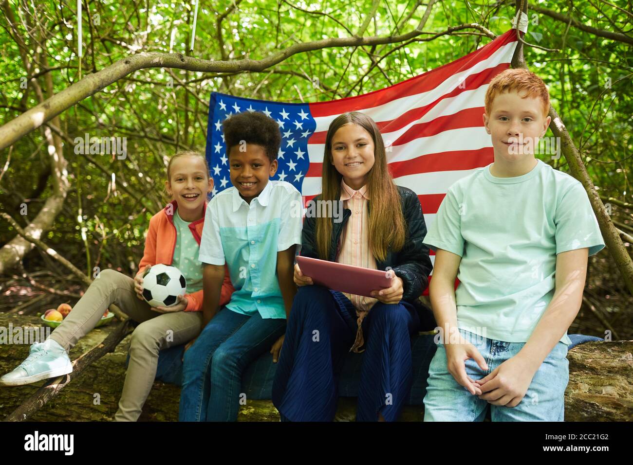 Groupe multiethnique d'enfants regardant la caméra tout en étant assis sous les branches d'un grand arbre et en jouant dans la forêt ou dans la cour arrière, espace de copie Banque D'Images