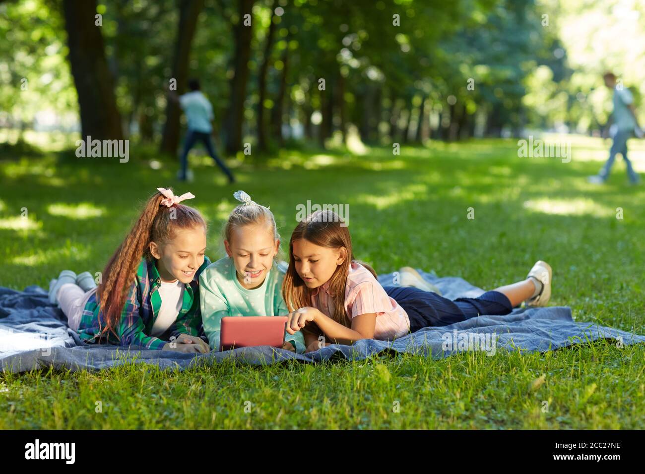 Portrait complet de trois adolescentes utilisant une tablette numérique tout en étant allongé sur de l'herbe verte dans le parc à l'extérieur éclairé par la lumière du soleil, espace de copie Banque D'Images