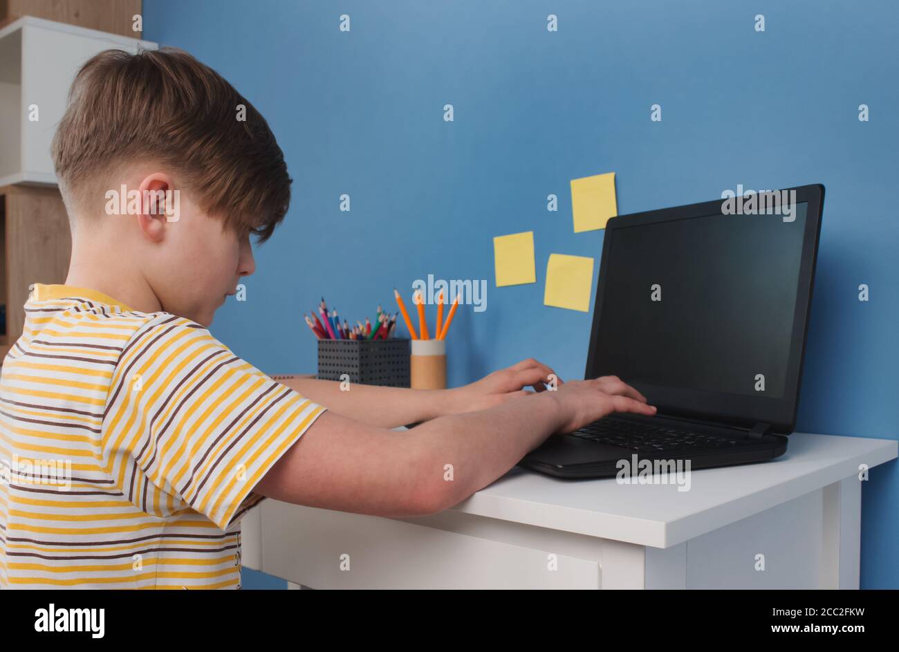 Garçon utilisant un ordinateur de bureau pour étudier en ligne pendant la quarantaine, assis à la table contre le mur bleu. Homeschooling, cours de formation en ligne ou Banque D'Images