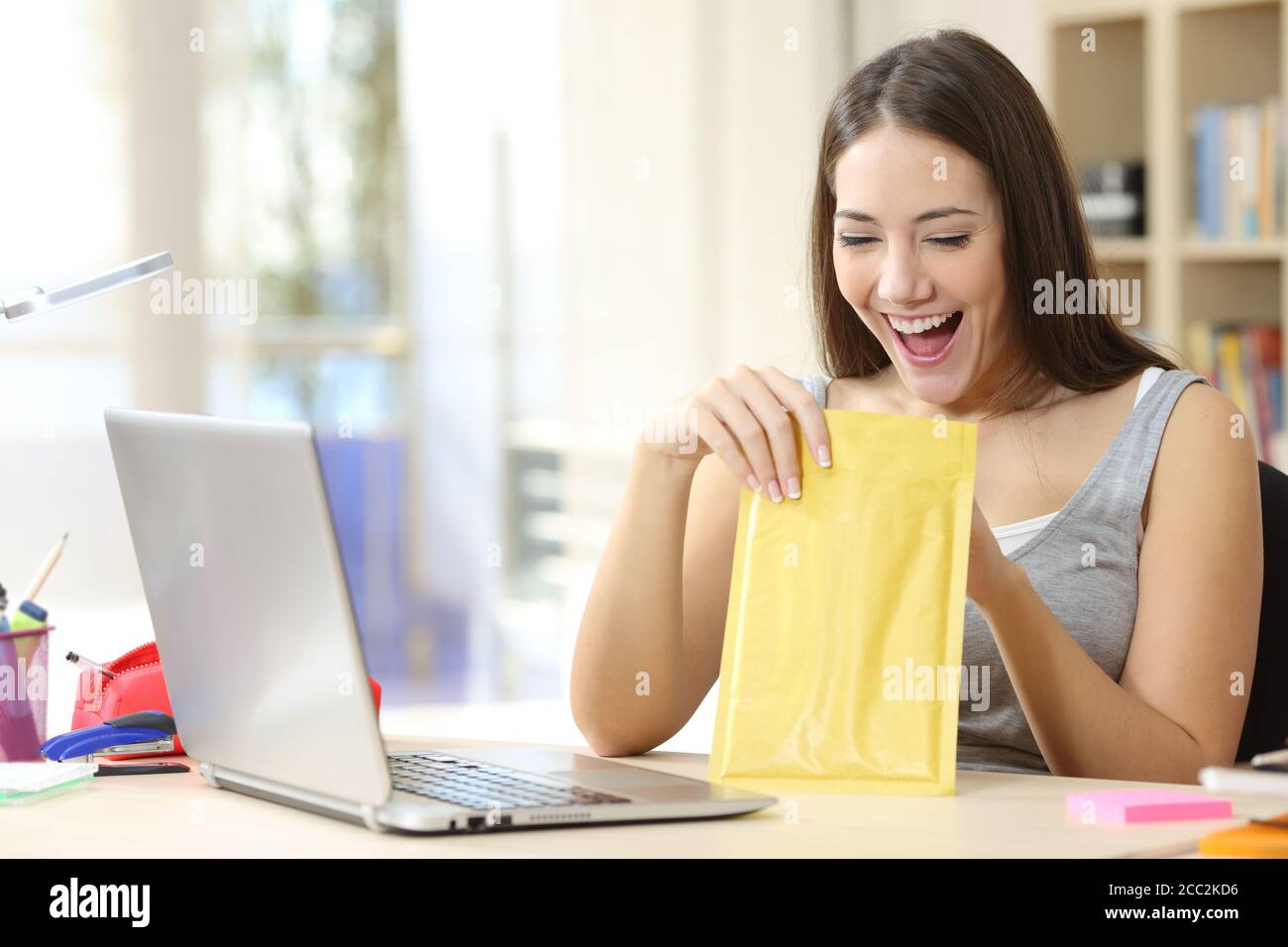 Une étudiante excitée ouvre et regarde à l'intérieur d'une enveloppe rembourrée assis sur un bureau à la maison Banque D'Images