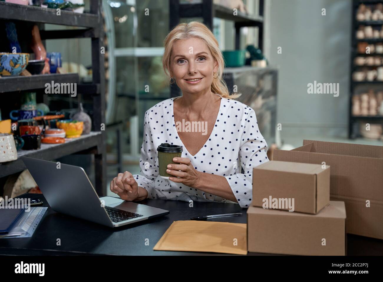 Café du matin. Portrait d'une belle femme d'affaires ou d'une femme d'affaires travaillant sur un ordinateur portable dans un studio d'art ou une boutique de poterie artisanale, buvant du café et souriant à l'appareil photo. Petite entreprise, vente au détail Banque D'Images