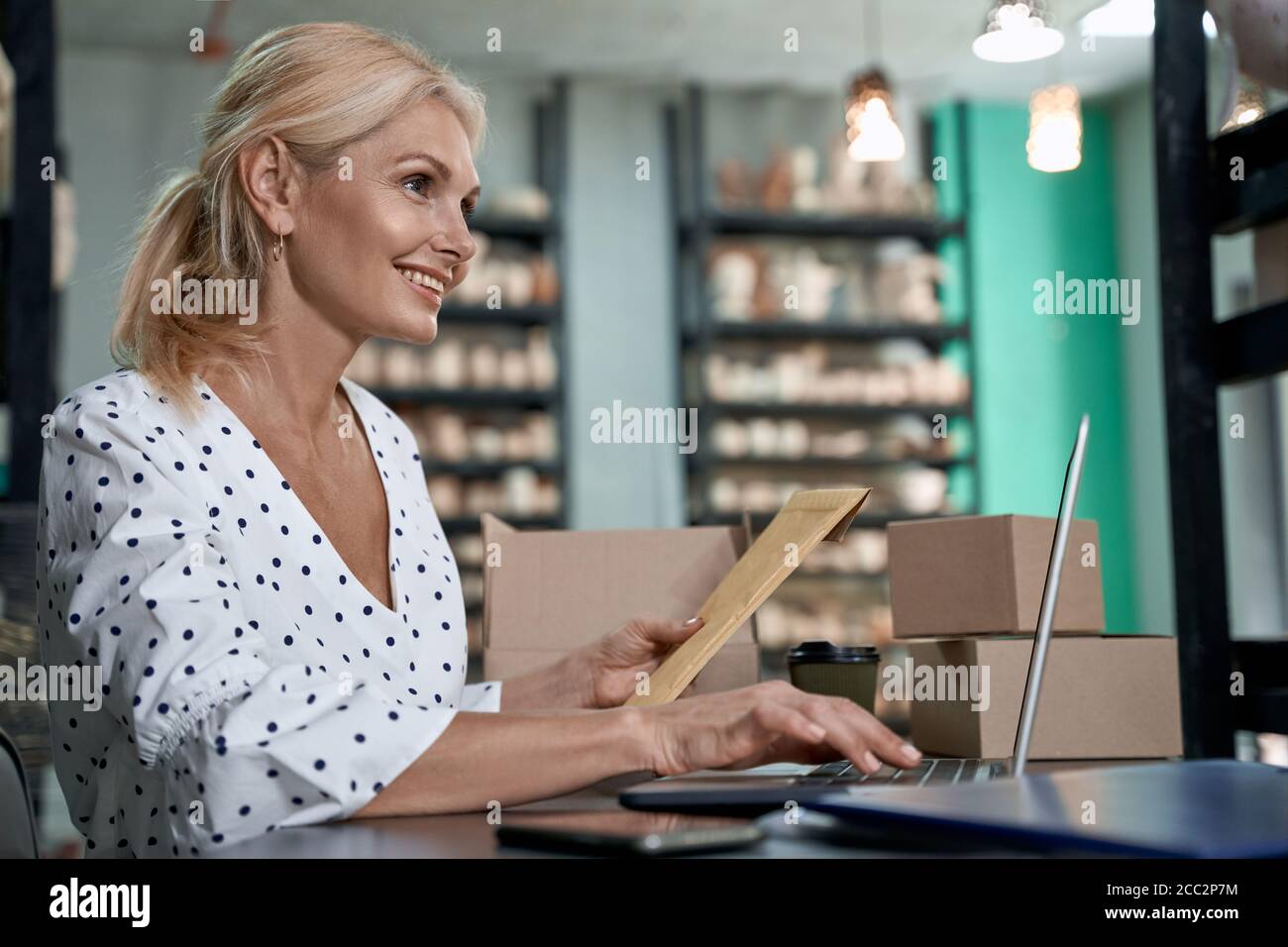 Vue latérale d'une heureuse réussite propriétaire d'affaires tenant une enveloppe ou un colis, en utilisant un ordinateur portable et souriant tout en étant assis dans sa boutique de poterie artisanale avec des produits céramiques faits à la main. Petite entreprise, vente au détail Banque D'Images