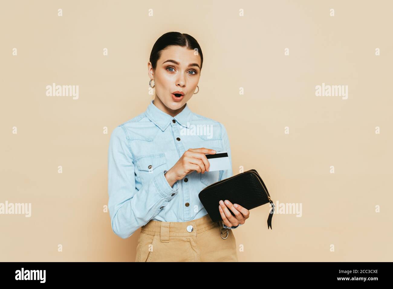 femme brune choquée dans une chemise en denim tenant un portefeuille et un crédit carte isolée sur beige Banque D'Images