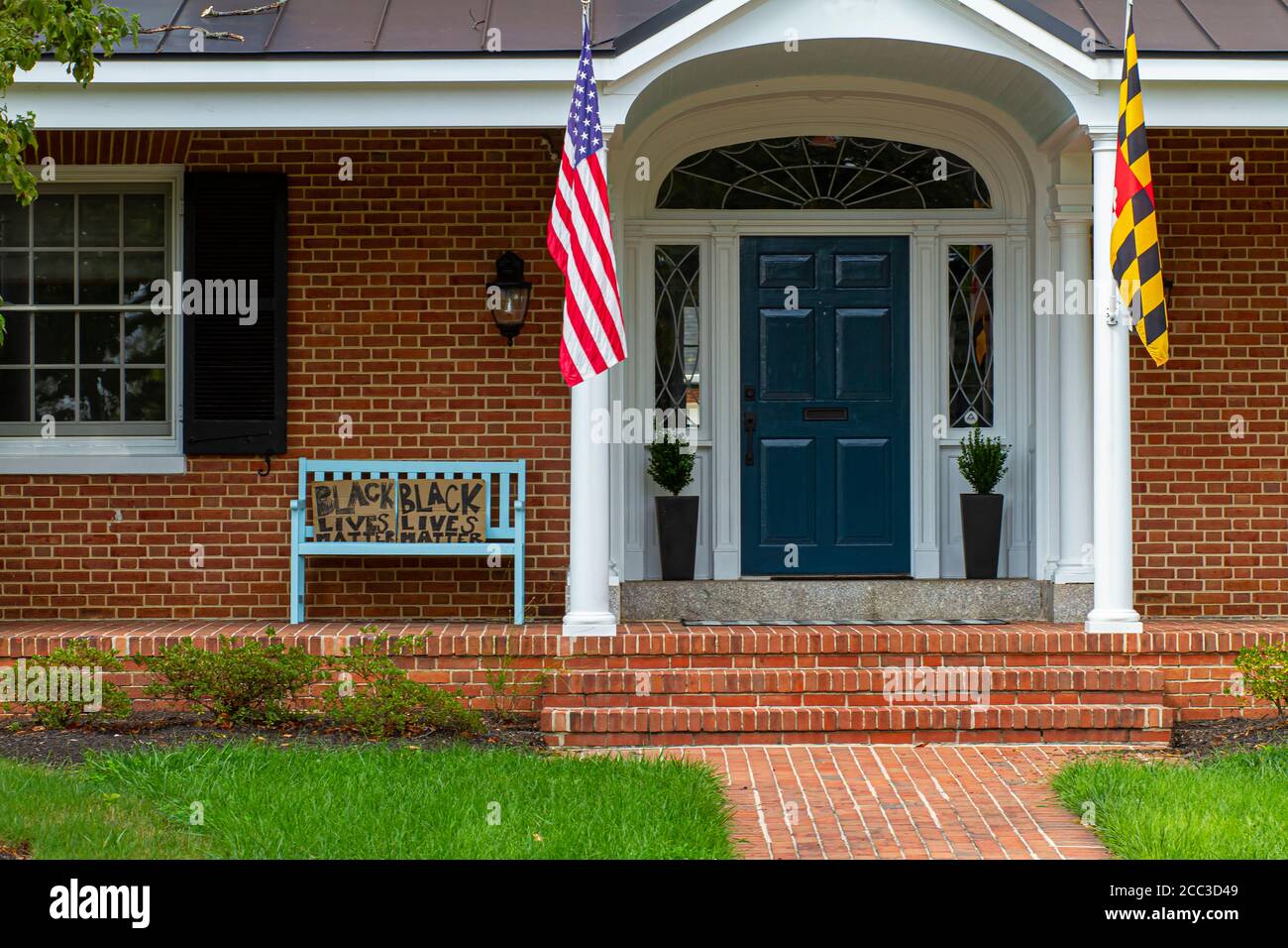 Frederick, MD 08/14/2020: Une maison traditionnelle en brique avec un drapeau américain et un drapeau du Maryland sur les poteaux de drapeau à l'entrée a un banc bleu vintage sur le Banque D'Images