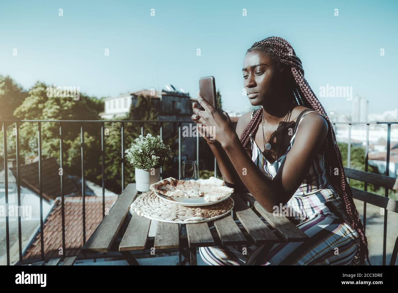 Une jeune femme noire pensive glamour utilise son smartphone dans un étui en cuir tout en étant assis à la table le balcon d'un restaurant en plein air Banque D'Images