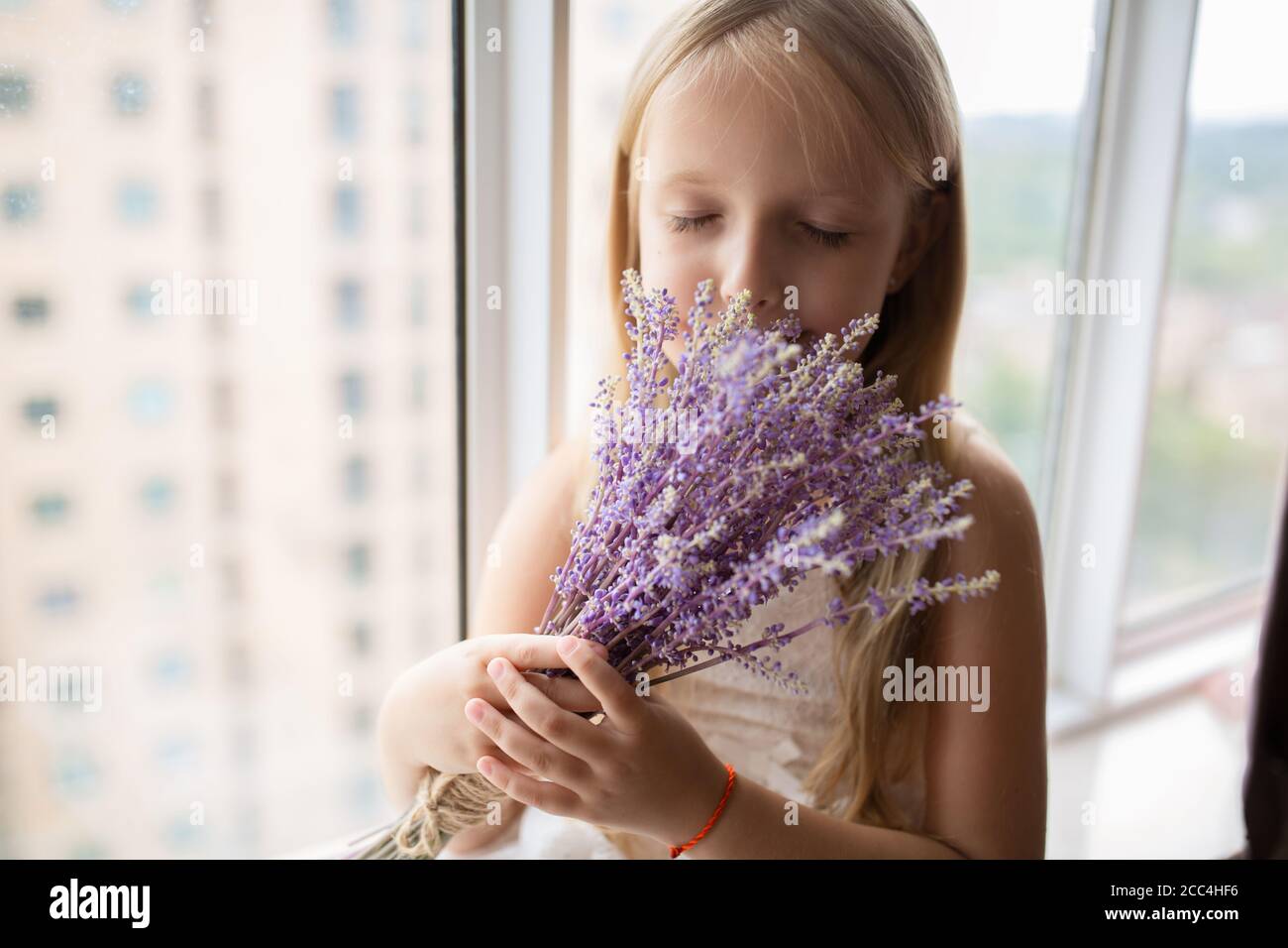 Jolie petite fille caucasienne avec cheveux blonds tenant un bouquet de fleurs violettes à la maison. Enfant debout près de la fenêtre pendant la pandémie de covid-19 du coronavirus Banque D'Images