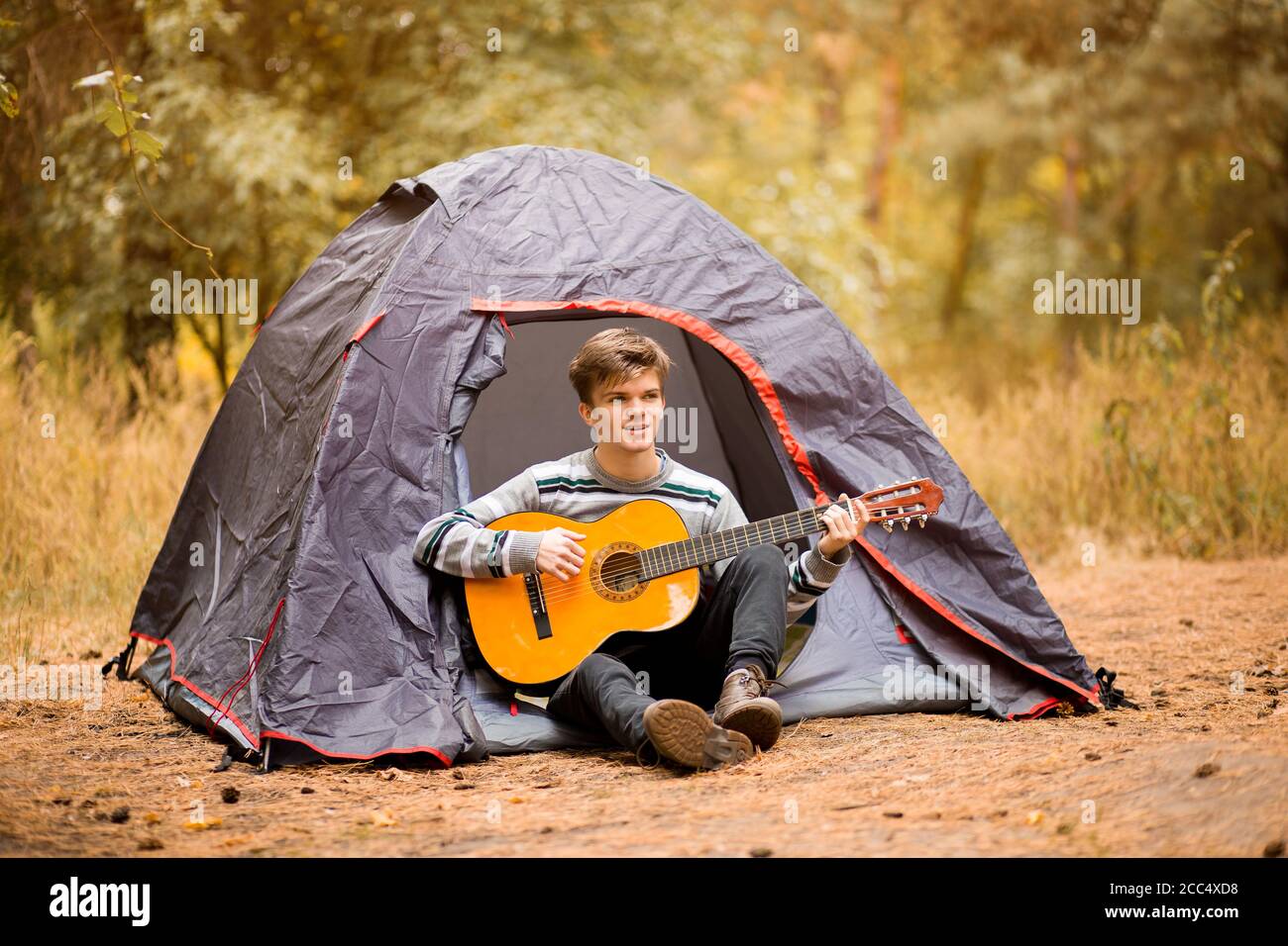 Jeune homme souriant assis près d'une tente touristique et jouant de la guitare dans la forêt d'automne Banque D'Images