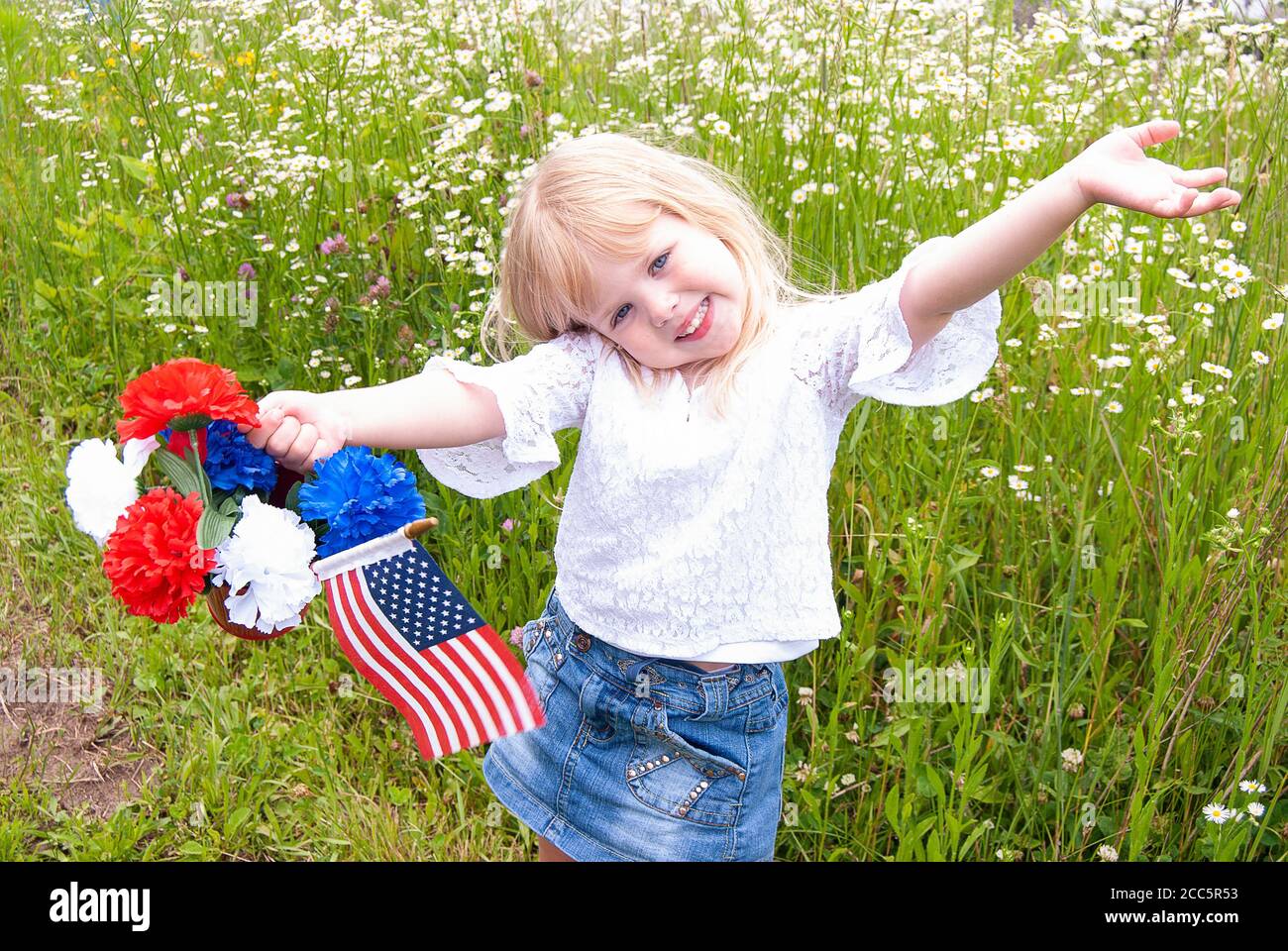Bonne petite fille blonde avec drapeau américain et bouquet de œillets dans le champ de fleurs sauvages Banque D'Images