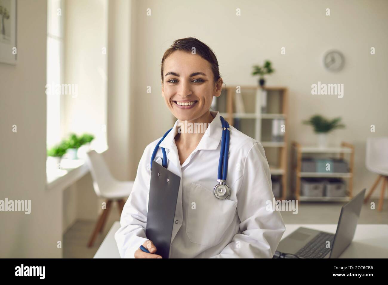 Jeune médecin thérapeute souriant debout et regardant la caméra plus bureau de la clinique médicale à l'arrière-plan Banque D'Images