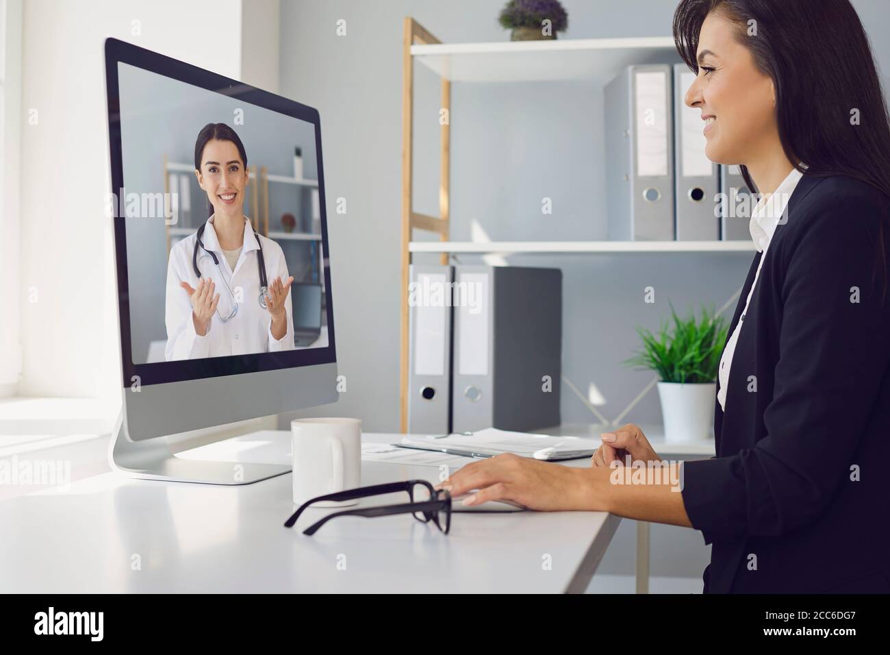 Jeune belle femme d'affaires est au bureau au travail utilise un appel vidéo pour parler à un médecin en ligne. Banque D'Images