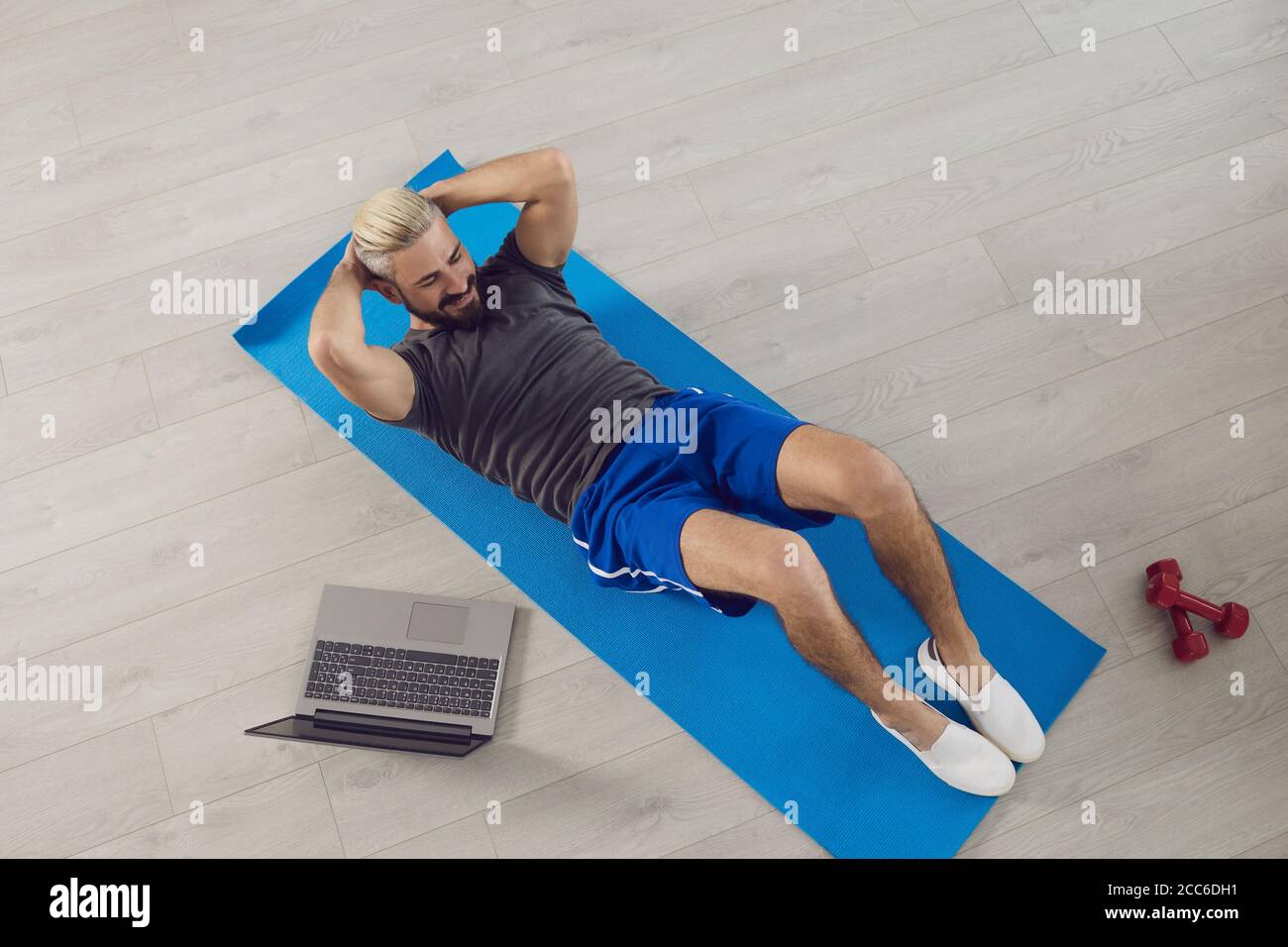 Un jeune homme en forme fait des exercices d'entraînement sur l'abs tout en étant allongé sur le sol, regarde un cours d'entraîneur en ligne tout en ayant un ordinateur portable dans la chambre. Banque D'Images