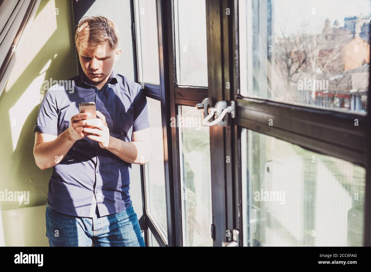 Jeune homme avec téléphone portable rester près de la fenêtre dedans lumière de jour contrastée Banque D'Images