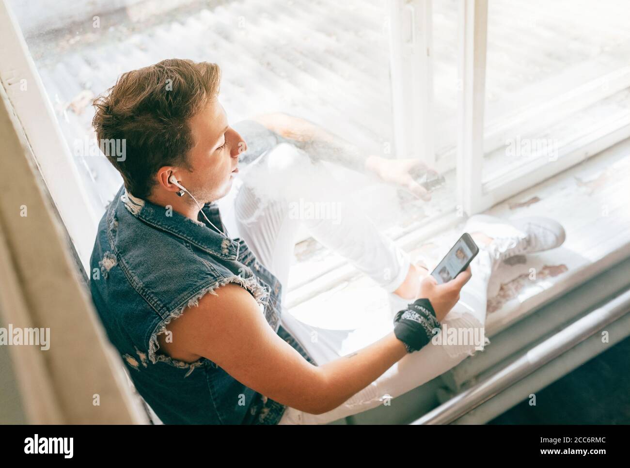 Portrait d'un adolescent moderne vêtu d'un Jean sans manches assis sur le rebord de la fenêtre, en écoutant la musique du téléphone à l'aide du casque et en fumant un électrique Banque D'Images