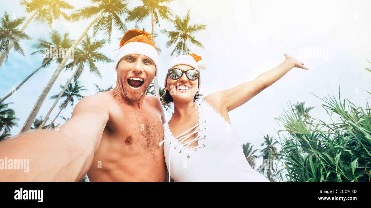 Image de Noël de jeunes gens gais couple habillé rouge chapeaux de Santa faisant selfie avec tropique palmiers fond . Banque D'Images