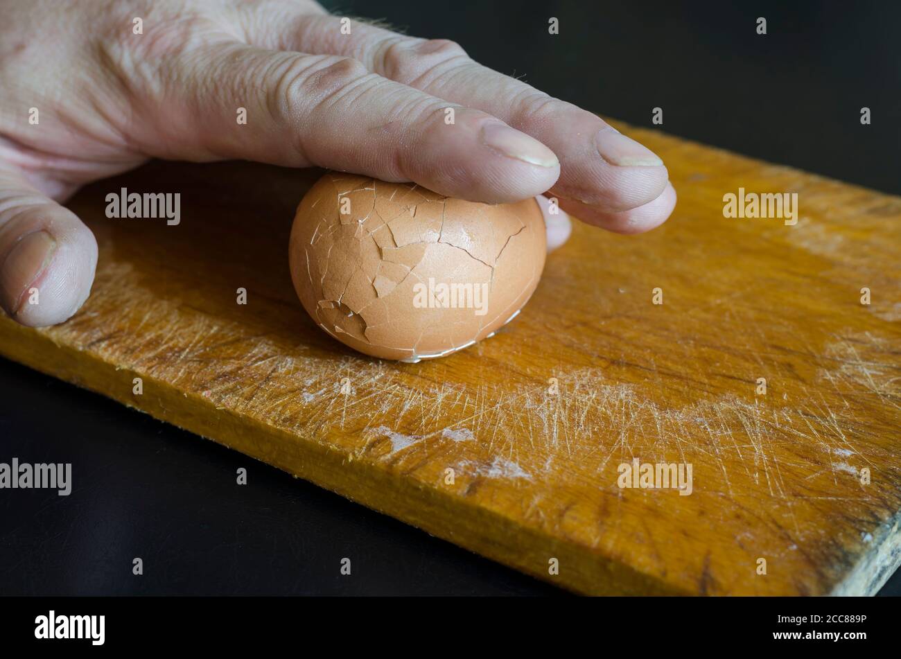 Une main mâle mature tient un œuf de poulet cuit dans sa coquille contre la planche à découper. Méthode de pelage des coquilles d'aubergines. Cuisson. Mise au point sélective. Banque D'Images