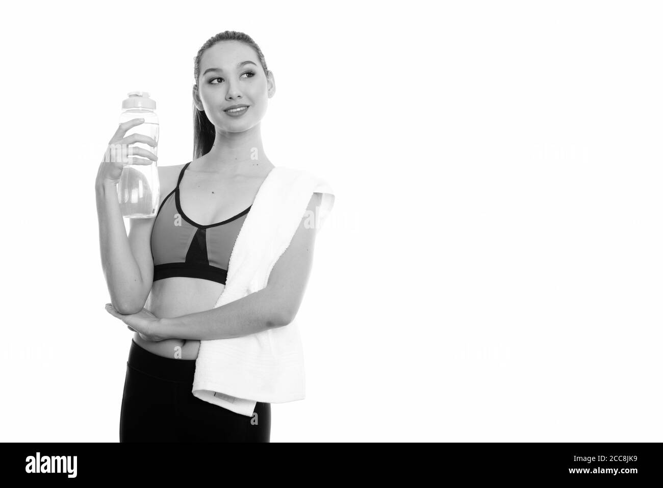 Studio shot of young happy Asian woman smiling et penser tout en tenant la bouteille de l'eau prêt pour salle de sport Banque D'Images