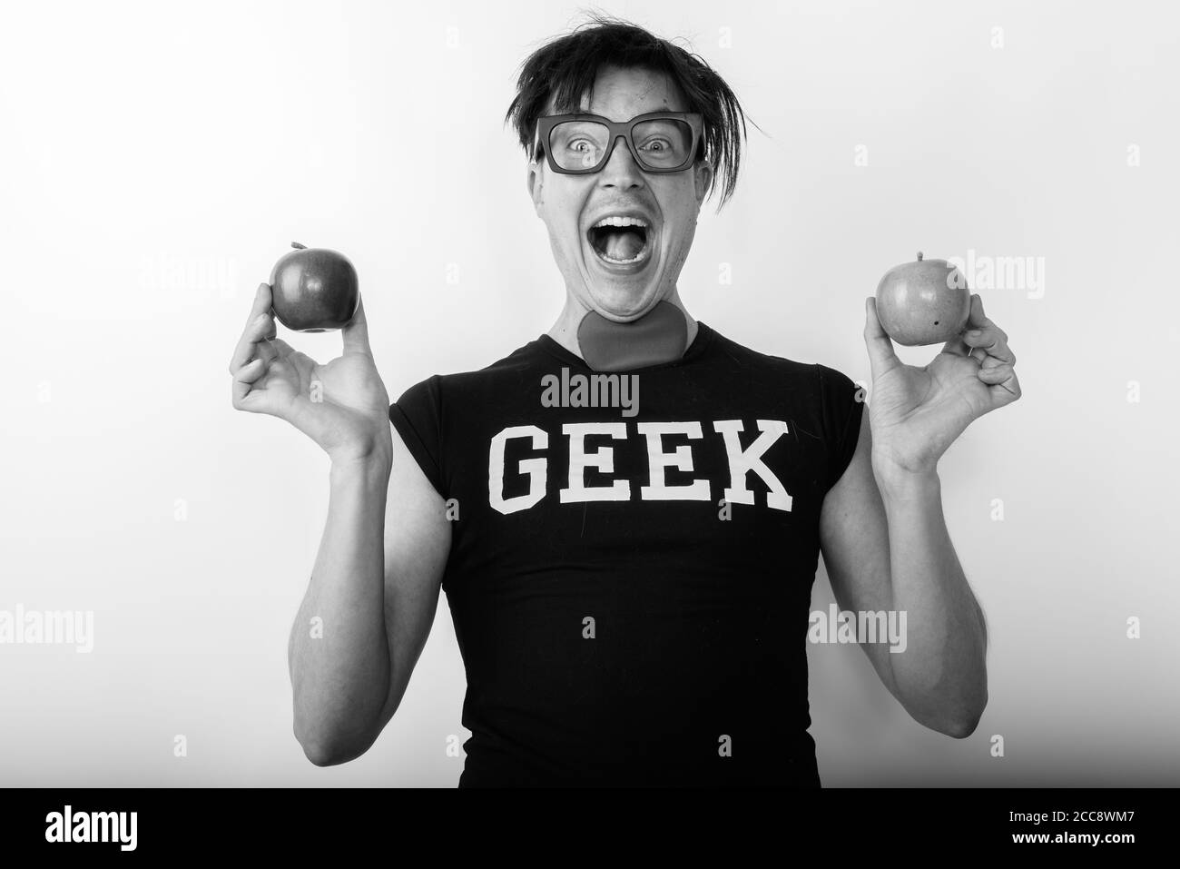 Photo studio d'un jeune homme nerd heureux souriant tout en tenant deux pommes avec coeur sur le cou Banque D'Images