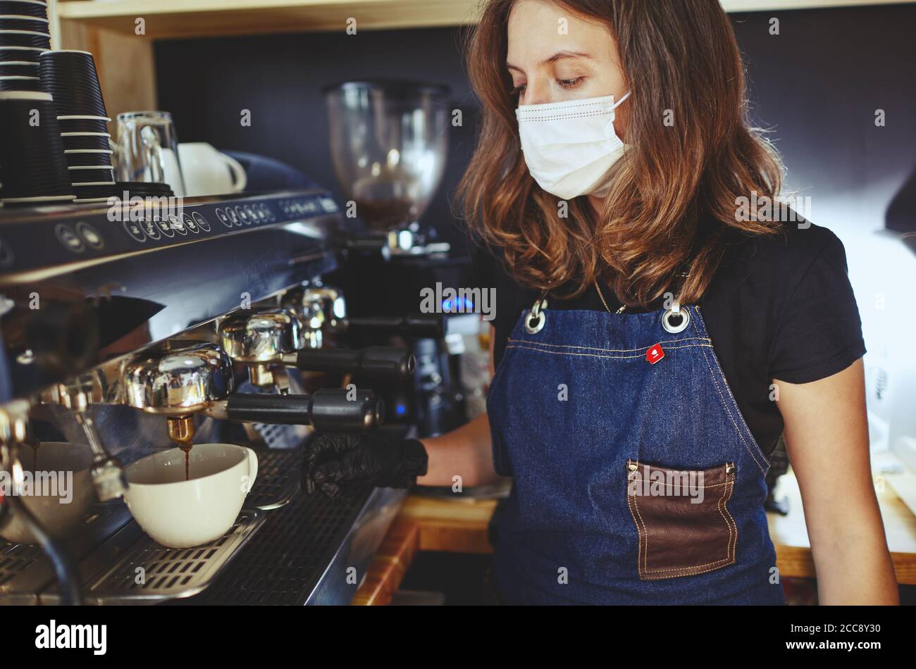 Barista portant un masque de protection du visage et des gants médicaux en latex noirs, préparant une boisson à base de café à l'aide d'une machine à espresso dans un café. Banque D'Images