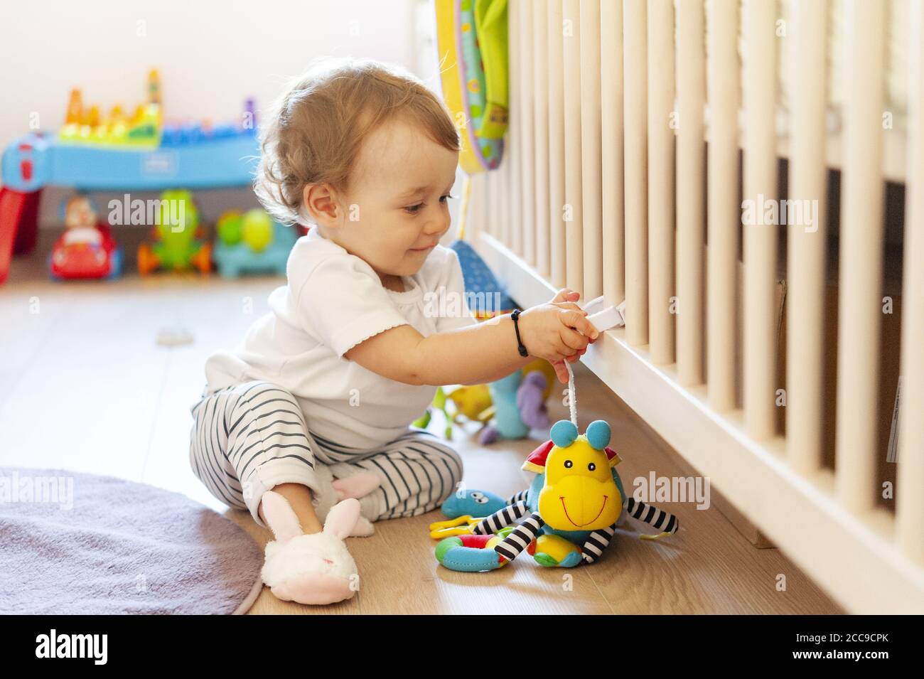 Petite fille souriante (16 mois) assise sur le sol et jouant dans sa chambre avec son jouet mignon préféré près de son berceau en bois blanc. reproduction ir Banque D'Images