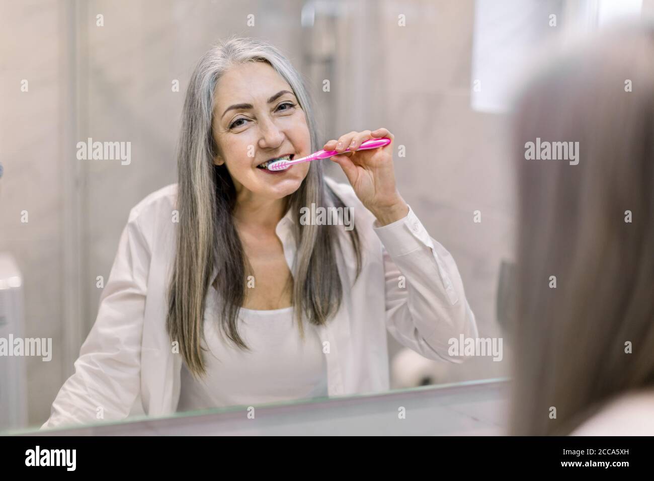 Reflet dans le miroir de la belle femme âgée souriante avec de longs cheveux gris droits, debout dans la salle de bains, se brossant les dents et appréciant son matin Banque D'Images