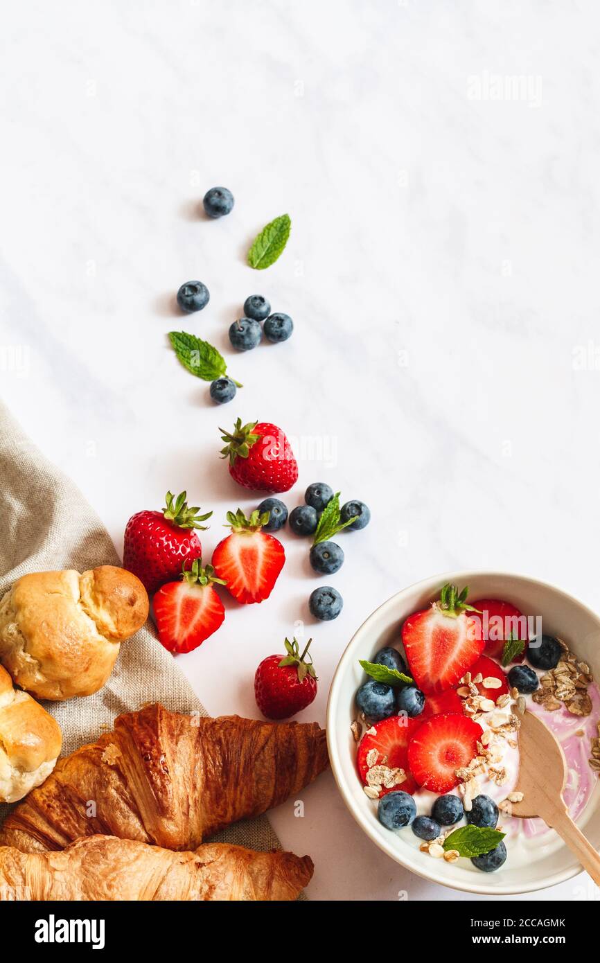 Petit déjeuner en été. Bol de yaourt avec fraises, bleuets et muesli, croissants et brioche parisienne sur fond de marbre blanc. Banque D'Images